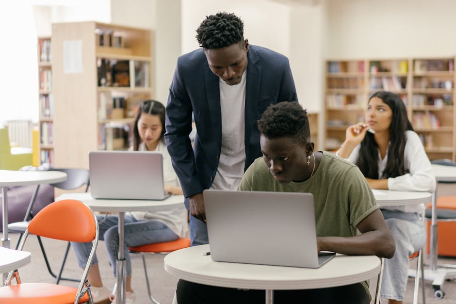 Students learning in a classroom setting with a teacher assisting and laptops on desks